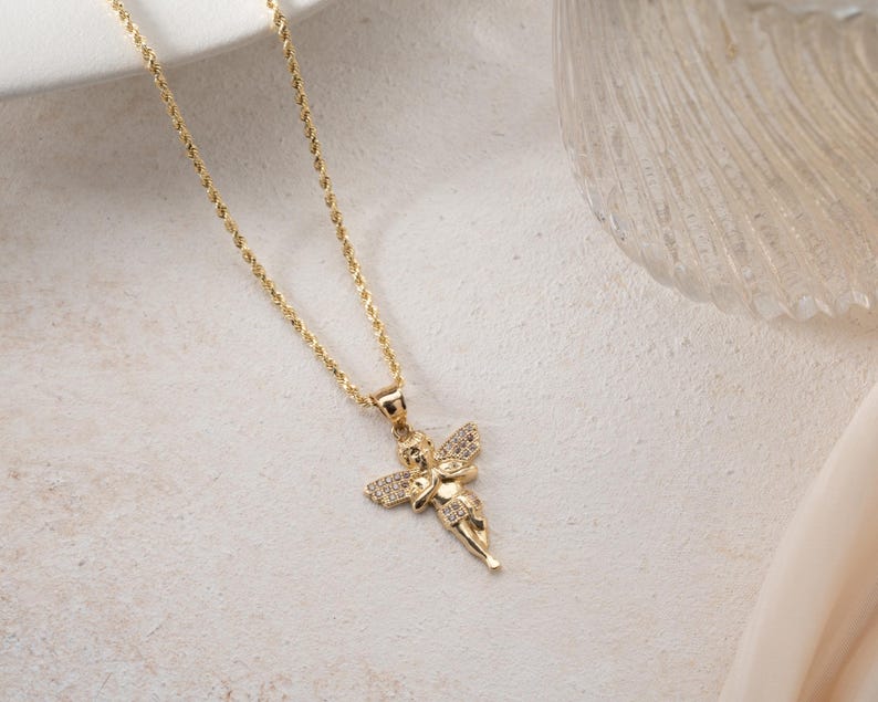 May include: A gold-toned rope chain necklace featuring a pendant shaped like a cherub with outstretched wings. The cherub figure is embellished with small, clear, sparkling stones set along the wings and body, creating a detailed metallic accessory.