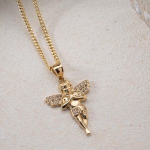 May include: A gold-toned necklace featuring a pendant shaped like a cherub with folded hands. The cherub's wings and lower garment are embellished with small, clear, sparkling stones. The pendant hangs from a delicate gold-toned curb chain.
