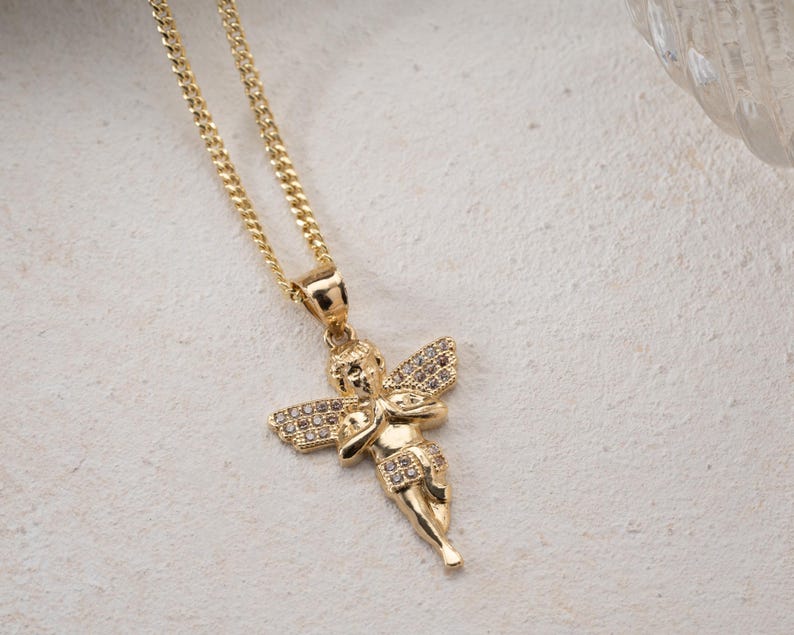 May include: A gold-toned necklace featuring a pendant shaped like a cherub with folded hands. The cherub's wings and lower garment are embellished with small, clear, sparkling stones. The pendant hangs from a delicate gold-toned curb chain.