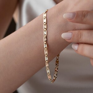 Puede incluir: Una pulsera de cadena dorada que presenta una serie de eslabones planos de forma ovalada. Cada eslab&oacute;n est&aacute; pulido con un patr&oacute;n texturizado en forma de estrella en el centro, creando un dise&ntilde;o repetitivo que capta la luz a lo largo de la joya.