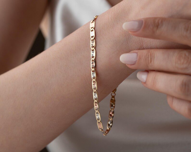 Puede incluir: Una pulsera de cadena dorada que presenta una serie de eslabones planos de forma ovalada. Cada eslab&oacute;n est&aacute; pulido con un patr&oacute;n texturizado en forma de estrella en el centro, creando un dise&ntilde;o repetitivo que capta la luz a lo largo de la joya.