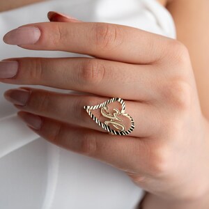 Op de afbeelding: Een goudkleurige ring in de vorm van een hart, met een gestileerde letter "M" in het midden. De omtrek van het hart heeft een textuur, touwachtig ontwerp. De ring wordt aan een vinger gedragen.