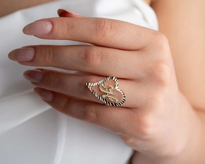 Op de afbeelding: Een goudkleurige ring in de vorm van een hart, met een gestileerde letter "M" in het midden. De omtrek van het hart heeft een textuur, touwachtig ontwerp. De ring wordt aan een vinger gedragen.