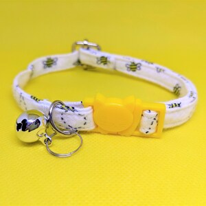 May include: White cat collar with a yellow bee design and a silver bell. The collar has a yellow plastic buckle.