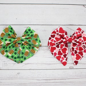 Può includere: Due fiocchi per capelli in tessuto. Uno è verde con boccali di birra e trifogli. L'altro è rosso con vari motivi a cuore. Entrambi i fiocchi sono su una superficie di legno bianco.