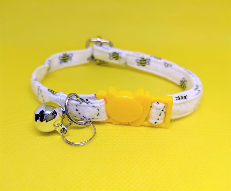May include: White cat collar with a yellow bee design and a silver bell. The collar has a yellow plastic buckle.