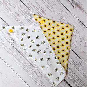 May include: A reversible white bandana with a yellow bee and sunflower print. One side features a pattern of yellow bees flying around a white background. The other side features a pattern of yellow sunflowers on a white background. The bandana has yellow snaps.