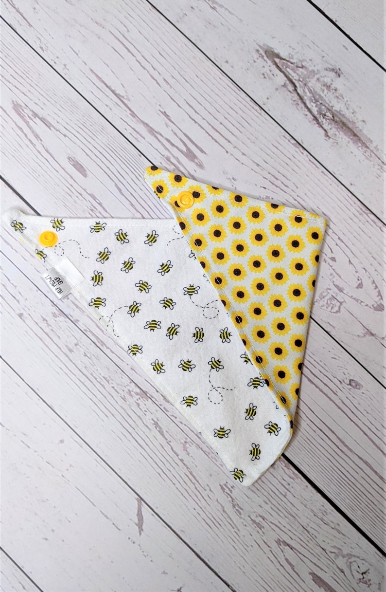May include: A reversible white bandana with a yellow bee and sunflower print. One side features a pattern of yellow bees flying around a white background. The other side features a pattern of yellow sunflowers on a white background. The bandana has yellow snaps.