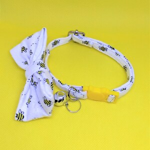 May include: White cat collar with a bow tie and a bell. The collar and bow tie have a pattern of black and yellow bees. The collar has a yellow plastic buckle.