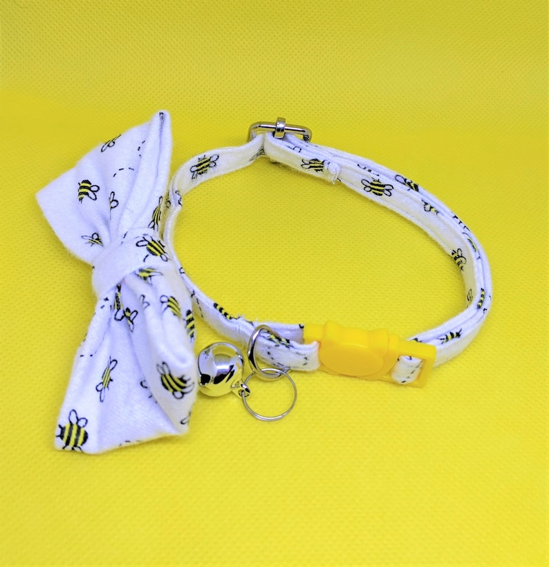 May include: White cat collar with a bow tie and a bell. The collar and bow tie have a pattern of black and yellow bees. The collar has a yellow plastic buckle.