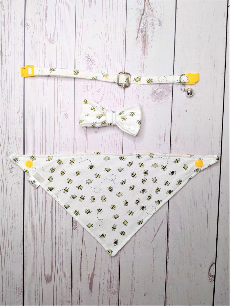 May include: A white pet bandana with a yellow bee pattern, a white bow tie with a yellow bee pattern, and a white collar with a yellow bee pattern and a yellow bell.