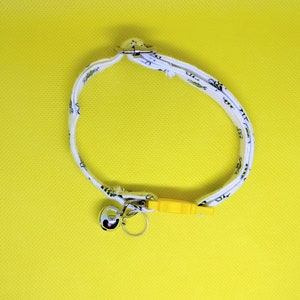 May include: White cat collar with a black and white pattern, a silver bell, and a yellow plastic safety buckle.