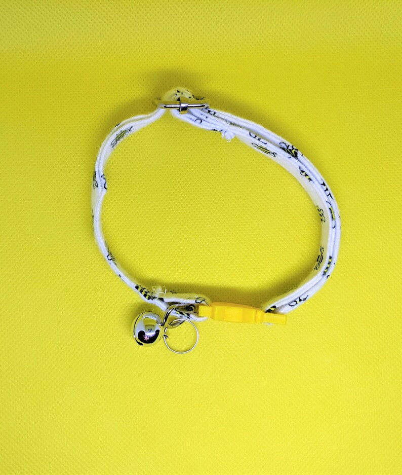 May include: White cat collar with a black and white pattern, a silver bell, and a yellow plastic safety buckle.