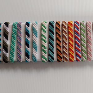 Puede incluir: Una colección de pulseras de cuentas coloridas dispuestas sobre una superficie blanca. Las pulseras presentan varias combinaciones de colores, incluyendo rosa, naranja, azul, verde y blanco, con un diseño retorcido. Las pulseras se exhiben en fila.