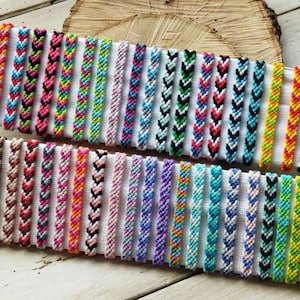 Può includere: Una collezione di braccialetti dell'amicizia colorati con motivi a chevron. I braccialetti sono esposti su una superficie bianca.