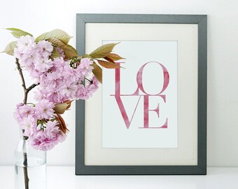 May include: A framed print with the word "LOVE" in a watercolor style, in shades of red. A clear glass vase holds a branch of pink cherry blossoms, adding a touch of nature to the composition. The frame is dark gray.