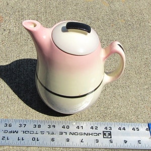 Peut inclure: Une théière en céramique rose et blanche avec une poignée et un couvercle noirs. La théière a une bande noire autour du milieu.