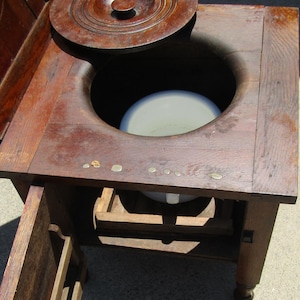 Puede incluir: Cómoda antigua de madera con tapa con bisagras y una pequeña puerta. La cómoda es de madera oscura y tiene una abertura redonda en el centro. La tapa está parcialmente abierta, revelando el interior. Es un mueble vintage.