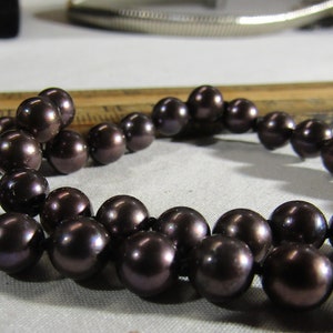 Może przedstawiać: Sznur ciemnobrązowych pereł, nawleczonych na cienki sznurek. Perły są okrągłe i gładkie, z lekkim opalizującym połyskiem.