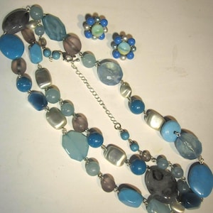 May include: A blue beaded necklace and matching earrings set. The necklace features a variety of blue, gray, and silver-toned beads in different shapes and sizes. The earrings are small, round, and have blue and silver accents.