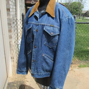 May include: A blue denim jacket with a brown corduroy collar. The jacket has two chest pockets with button closures and two lower hand pockets. The Wrangler jacket is displayed on a mannequin.