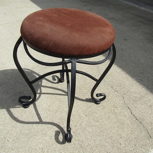Puede incluir: Un taburete redondo tapizado marrón con una base de hierro forjado negro. El taburete tiene un asiento circular y patas curvas con adornos decorativos. El asiento parece estar hecho de un material suave y texturizado.