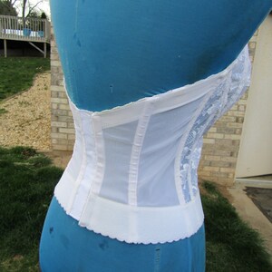 May include: White lace and mesh corset with a hook and eye closure in the back. The corset is on a blue dress form.