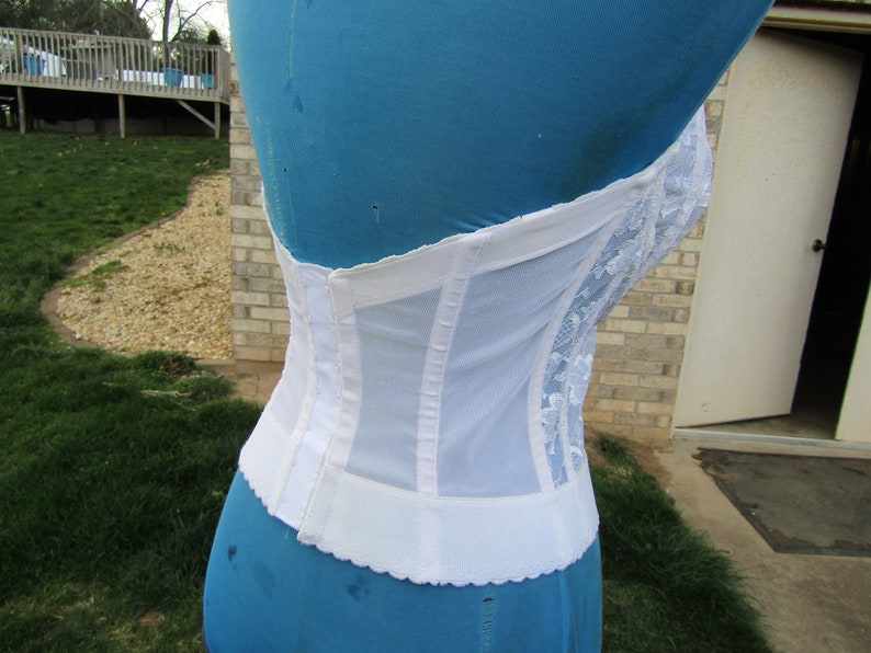 May include: White lace and mesh corset with a hook and eye closure in the back. The corset is on a blue dress form.