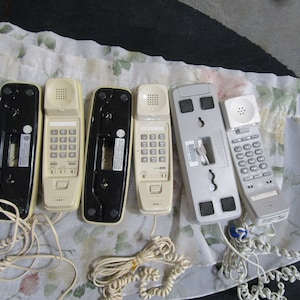 May include: Five vintage corded telephones in various colors, including black, white, and silver. The phones have a keypad and a speaker. The phones are on a white floral fabric background.
