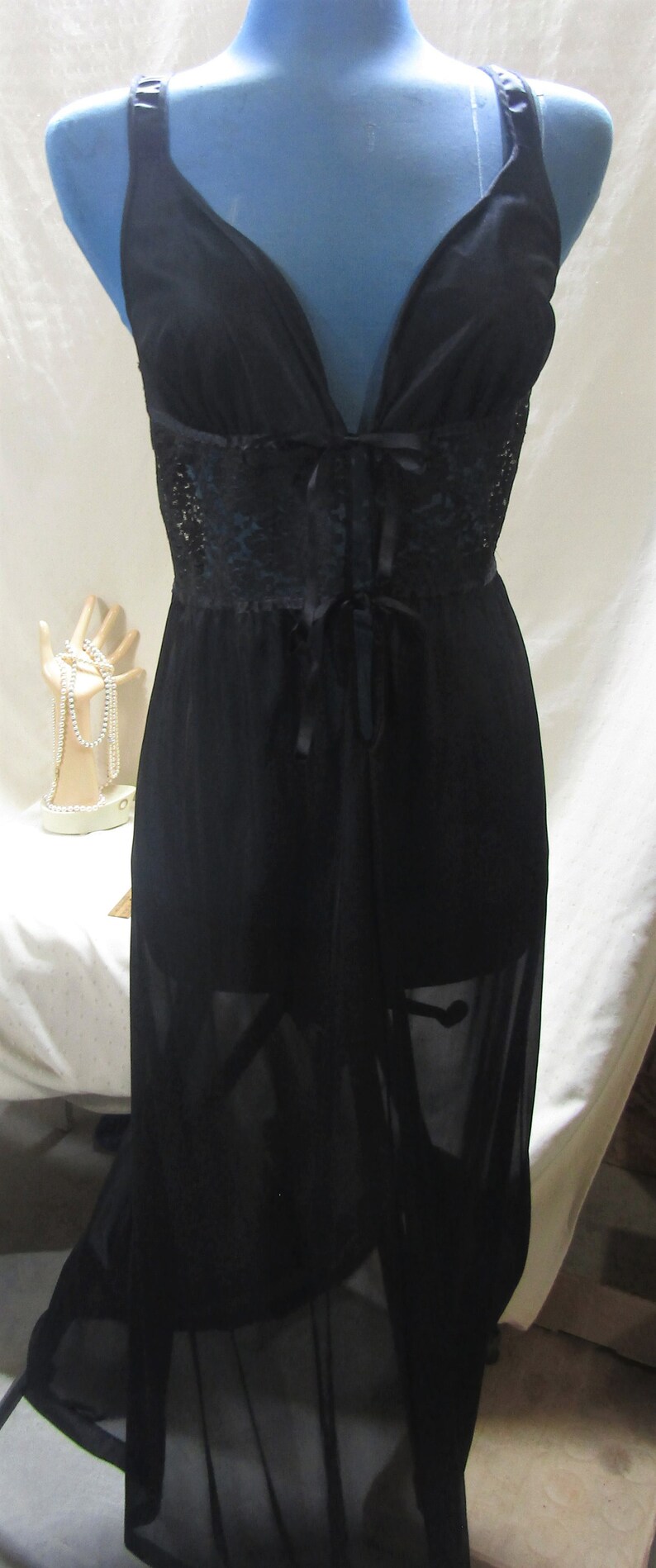 Pu&ograve; includere: Camicia da notte di lingerie nera con scollo a V profondo e dettagli in pizzo. L'abito presenta una gonna trasparente e una chiusura con fiocco in vita. L'indumento &egrave; esposto su un manichino blu.