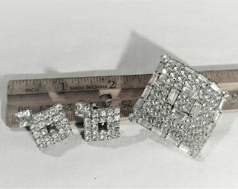 Può includere: Un set di tre gioielli in tono argento con strass. Il pezzo più grande è una spilla quadrata con un design geometrico. I due pezzi più piccoli sono orecchini quadrati.