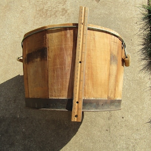 May include: A wooden bucket with a metal band around the top and a wooden handle. The bucket is sitting on a concrete surface with a ruler next to it.