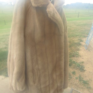 May include: A long, brown fur coat with a large collar and a single button closure. The coat is hanging on a white stand.