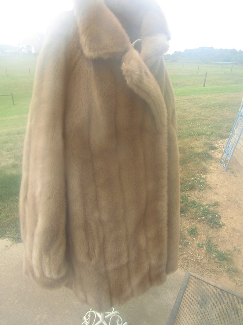 May include: A long, brown fur coat with a large collar and a single button closure. The coat is hanging on a white stand.