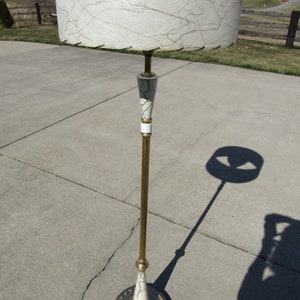 May include: A gold floor lamp with a white and gold patterned shade. The shade has a white fabric with gold lines and a gold trim. The lamp has a gold base with a white and gold patterned design.
