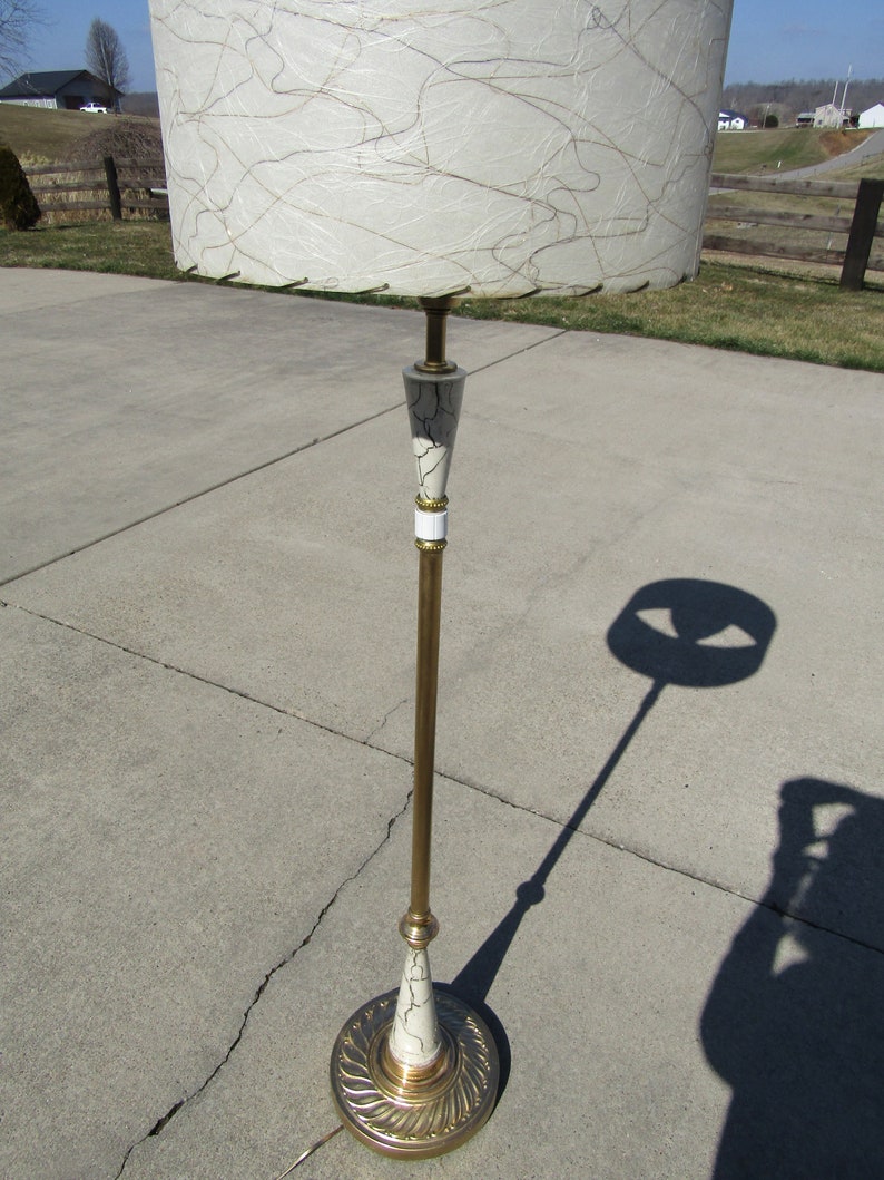 May include: A gold floor lamp with a white and gold patterned shade. The shade has a white fabric with gold lines and a gold trim. The lamp has a gold base with a white and gold patterned design.