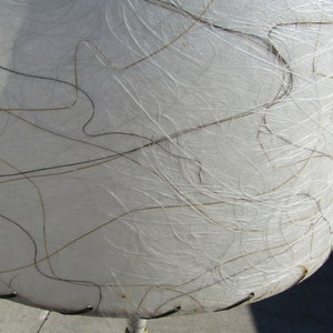 May include: White lampshade with a textured surface and brown lines. The lampshade is made of a material that resembles paper.