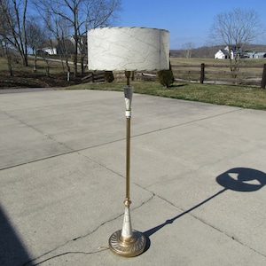 May include: A gold floor lamp with a white and gold patterned shade. The lamp has a tall, slender base and a round, decorative foot.