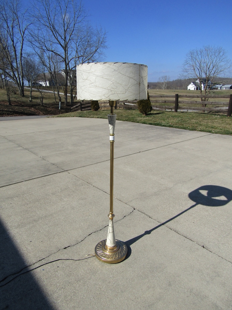 May include: A gold floor lamp with a white and gold patterned shade. The lamp has a tall, slender base and a round, decorative foot.