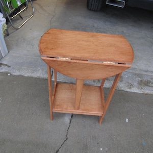 May include: A small, wooden drop-leaf table with a natural finish. The table has a rounded top that folds down, supported by a lower shelf and four legs. The table measures approximately 76 cm long when open.