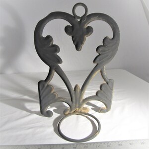 May include: Black metal wall-mounted plant hanger with a heart-shaped design and a ring at the bottom. The hanger has a distressed finish and intricate scrollwork.