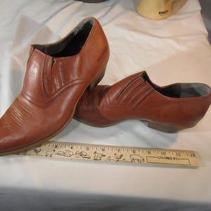 Könnte beinhalten: Ein Paar braune Cowboy-Stiefel aus Leder mit spitzer Zehenpartie und einem Pull-On-Design. Die Stiefel haben ein dekoratives Steppdetail am Schaft und ein kleines Etikett an der Seite mit der Aufschrift "DeLano West".