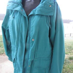 Può includere: Giacca color verde acqua con collo alto e cappuccio. La giacca presenta una cerniera a tutta lunghezza, due tasche anteriori e una vita con coulisse. La giacca è esposta su un manichino.