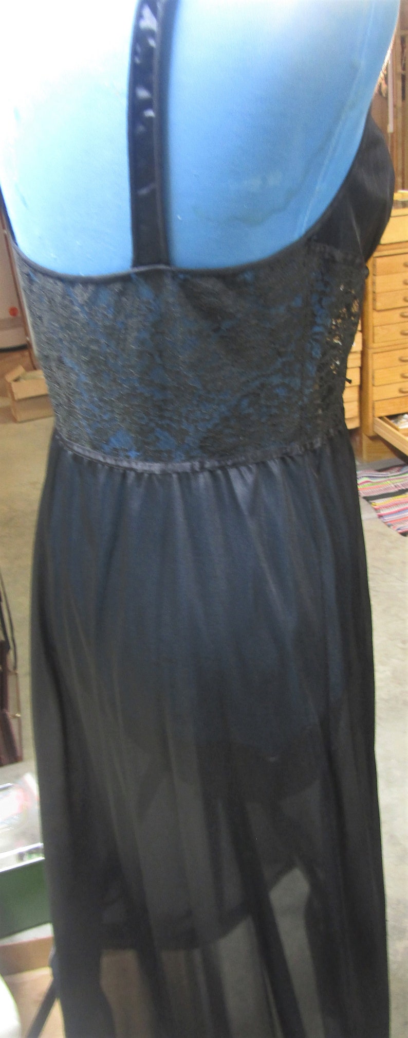 Pu&ograve; includere: Camicia da notte nera con corpetto in pizzo e spalline sottili. La gonna trasparente scende fino a terra. La camicia da notte &egrave; esposta su un manichino blu.
