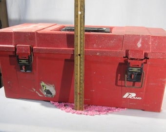 May include: Red plastic toolbox with black latches and a Plano logo. The toolbox is sitting on a white surface with a pink doily.