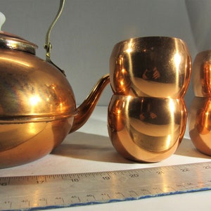 May include: A copper tea kettle with a white ceramic lid and a brass handle, next to a set of copper cups. The cups are stacked in pairs. A ruler is in the foreground.