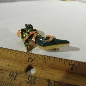 Puede incluir: Un broche en forma de zapato verde y dorado con detalles en rosa y verde. El broche mide aproximadamente 5 cm de largo y está sobre una regla de madera.