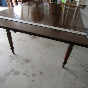 May include: A dark brown wooden table with a drop leaf extension. The table has turned legs with a rounded ball at the bottom. The table is sitting on a concrete floor.