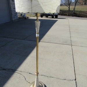 May include: A gold floor lamp with a white and gold patterned base and a white, textured lampshade. The lamp is standing on a concrete surface.