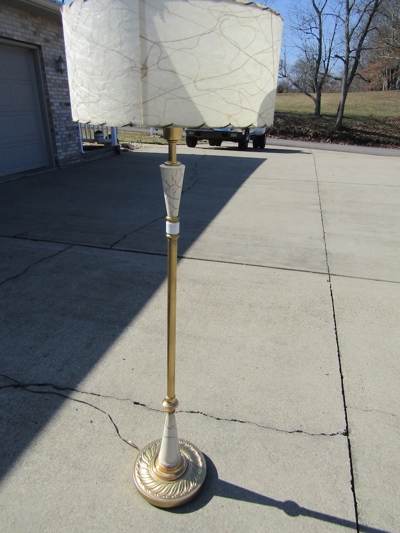 May include: A gold floor lamp with a white and gold patterned base and a white, textured lampshade. The lamp is standing on a concrete surface.